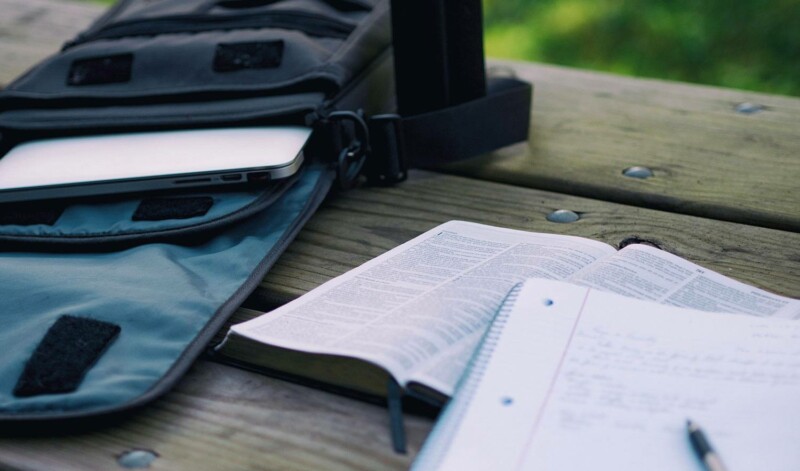 A student's backpack, laptop, textbook, and notebook on a park table for studying outside
